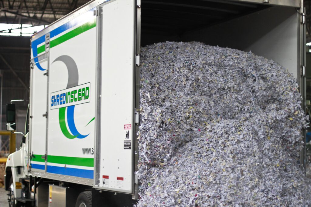 Photograph of a white box truck from "SHREDINSTEAD" with its rear door open, revealing a large pile of shredded paper. The paper is mostly white and grey with colorful flecks. The truck's side has a blue and green logo with the company name.