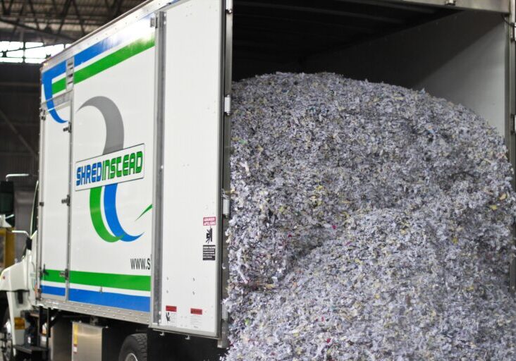 Photograph of a white box truck from "SHREDINSTEAD" with its rear door open, revealing a large pile of shredded paper. The paper is mostly white and grey with colorful flecks. The truck's side has a blue and green logo with the company name.